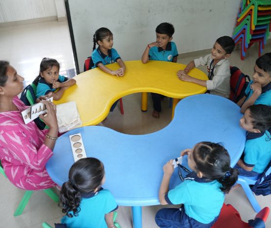 A female teacher helps a young girl with long hair and a blue bow as she writes in a notebook at a classroom desk, with other students visible in the background.